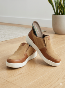 Tan slip-on sneakers with white chunky sole on a wooden floor; a potted plant in the background.