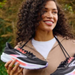 Woman with curly hair smiling while holding a pair of black and pink running shoes outdoors