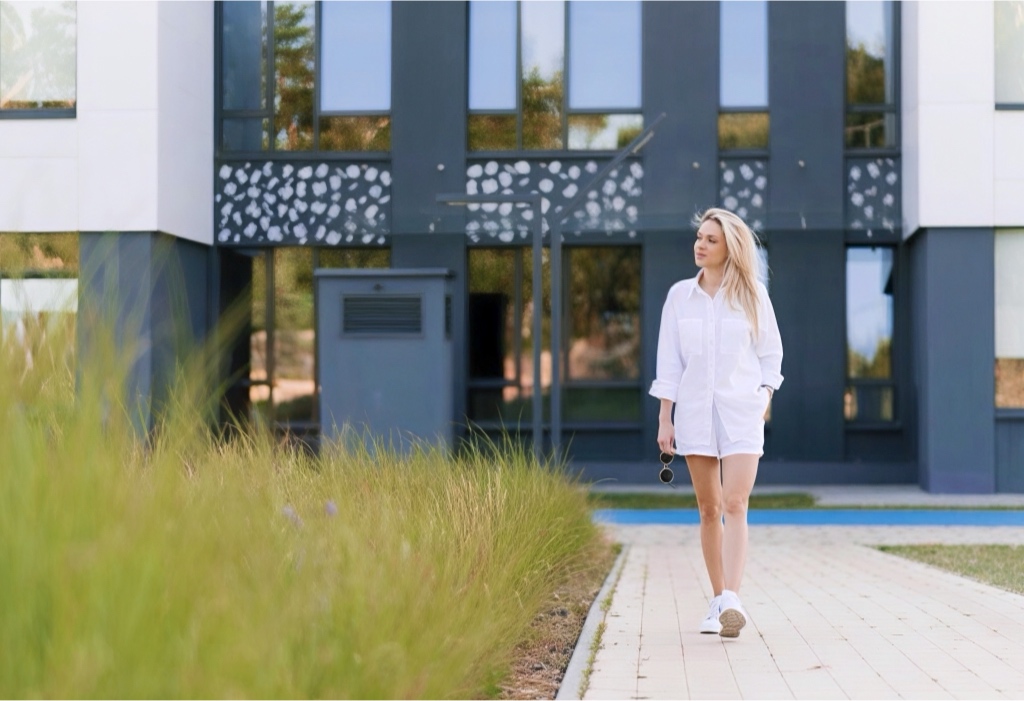 Woman in a white shirt and shorts walks along a paved campus path with a modern glass-and-stone building in the background.
