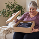 Older woman sits on a sofa, opening a shoebox containing a pair of sneakers with a smile on her face at home.