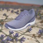 Bazaart_20260712_094701_810 - Rejuva Sole™ Lavender running shoe on sandy ground with purple flowers in the foreground.