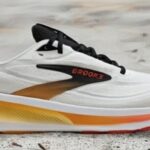 White Brooks running shoe with black laces and a yellow–orange gradient midsole on a sandy surface, side view.
