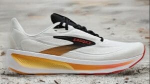 Bazaart_20260714_034044_661~2 - Rejuva Sole™ White Brooks running shoe with black laces and a yellow–orange gradient midsole on a sandy surface, side view.