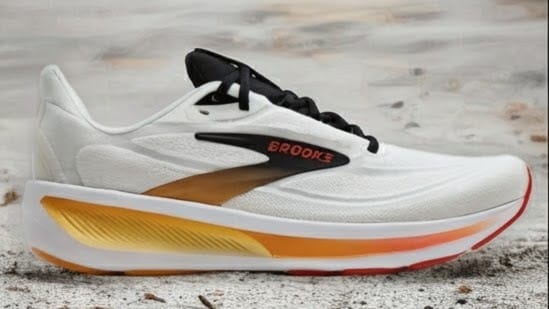 White Brooks running shoe with black laces and a yellow–orange gradient midsole on a sandy surface, side view.