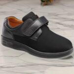 Black slip-on sneaker with a wide Velcro strap and orange interior resting on a marble floor.