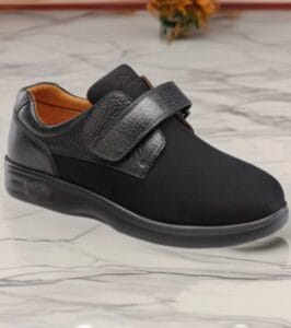 Black slip-on sneaker with a wide Velcro strap and orange interior resting on a marble floor.