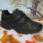 Black sneakers with two Velcro straps on a white surface, autumn leaves scattered around and a blurred fall backdrop behind them.
