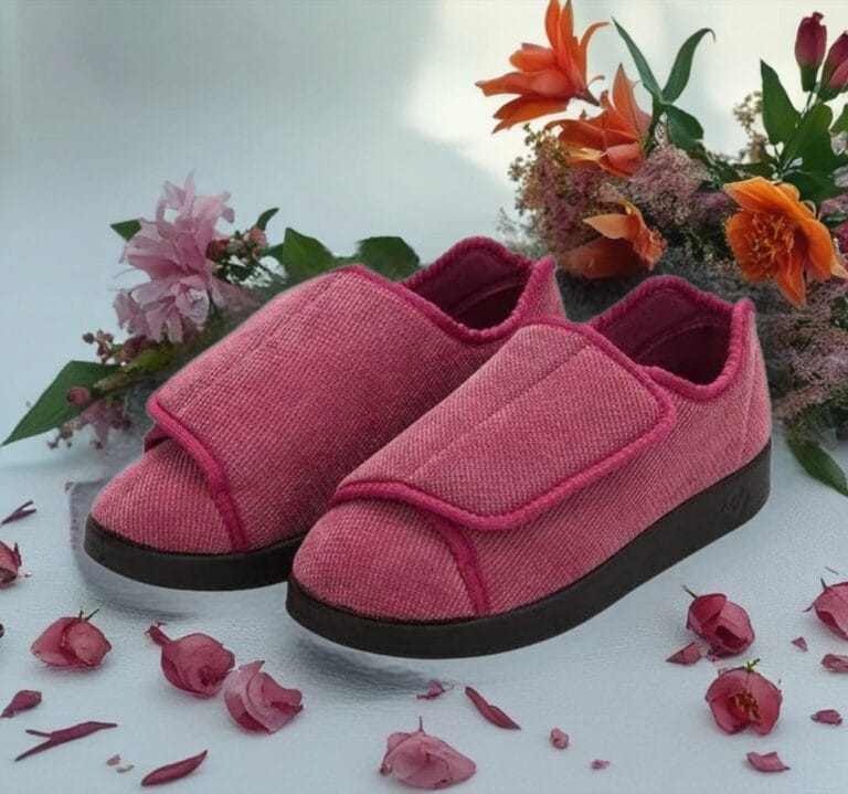 Pink fabric slip-on slippers with a Velcro strap and black sole, shown on a light surface among pink and orange flowers.