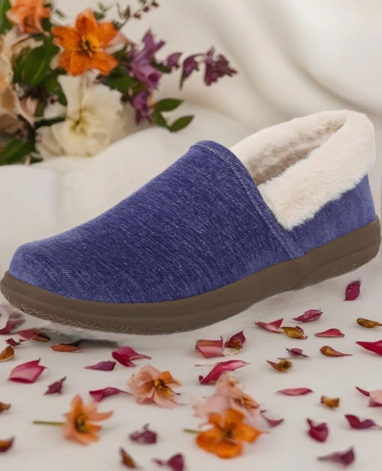 Blue denim slip-on slipper with plush white lining and brown sole, photographed on a white surface with flower petals in the foreground and background flowers.