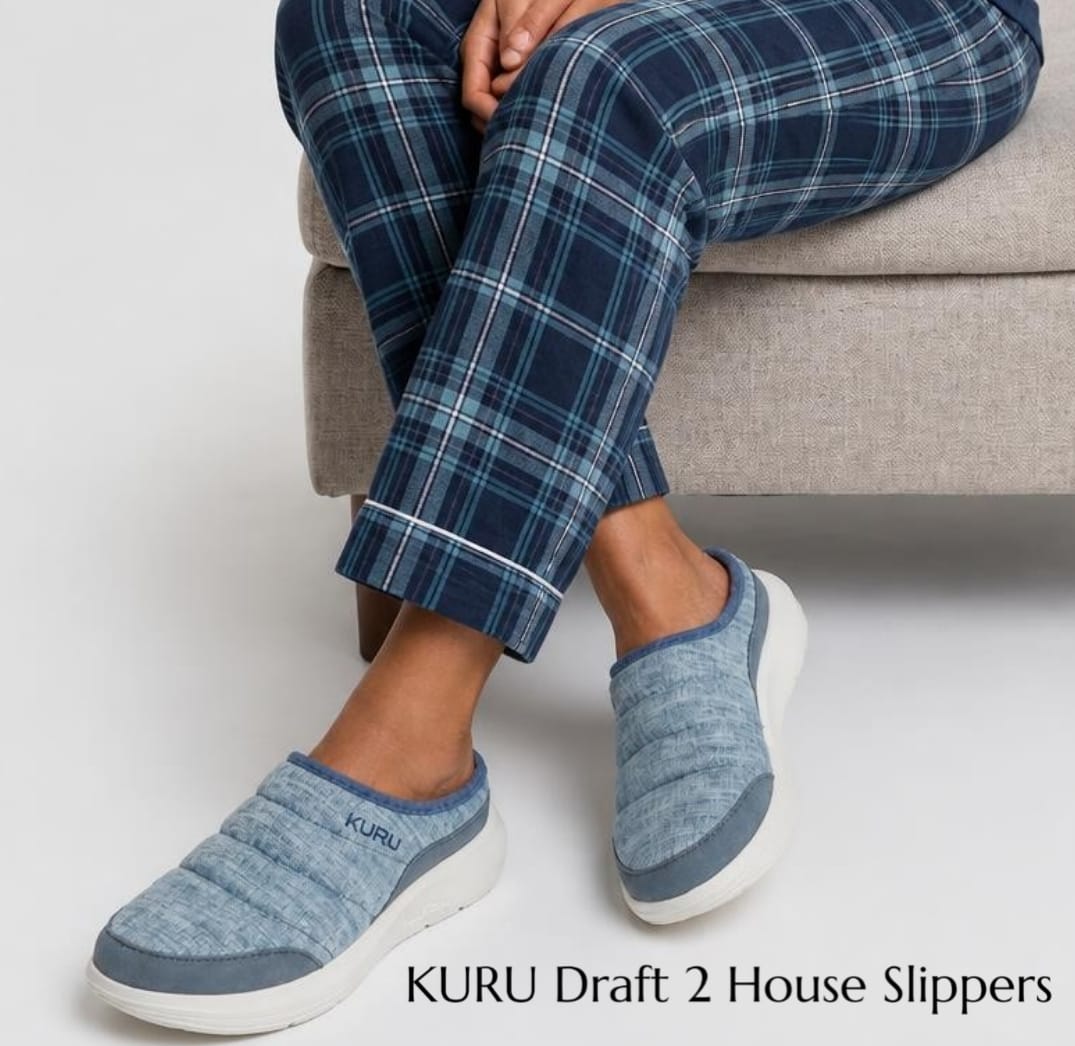 Person seated wearing blue plaid pajama pants and KURU Draft 2 blue house slippers, showcasing indoor footwear.